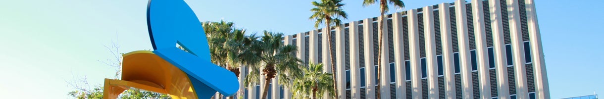 A tall building is the background. In the forefront is a blue and yellow sculpture. In between the art and the building are palm trees.