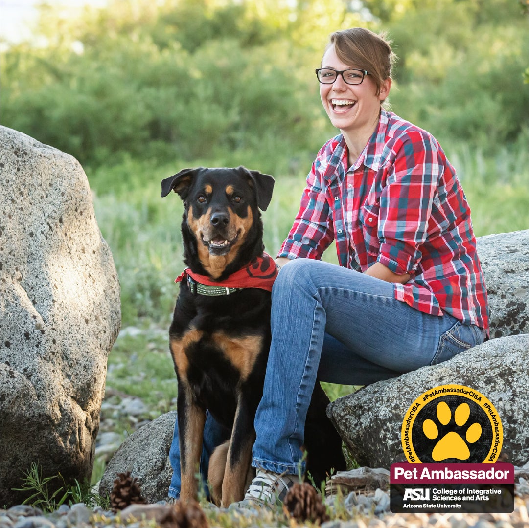 Max the rottweiler is sitting in field between two rocks. On the rock to his left, sits his human Mom Mara. She has her arm around him. Max, who is wearing a red bandana, and Mara, who is wearing red flannel, are happy and you can tell these two are best friends.