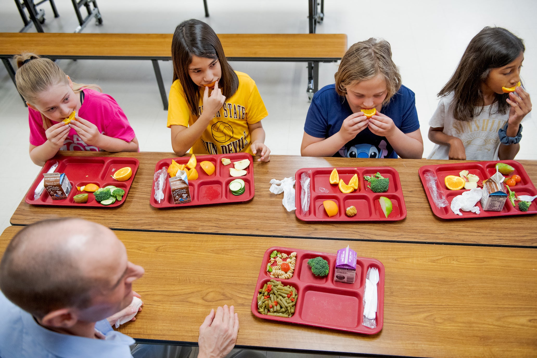 Overhead view of 4 children and one adult man sitting at a school cafeteria table. Each of the people have a red lunch tray containing fruits and vegetables sitting in front of them on the table. Some of the children are eating the fruits and vegetables.