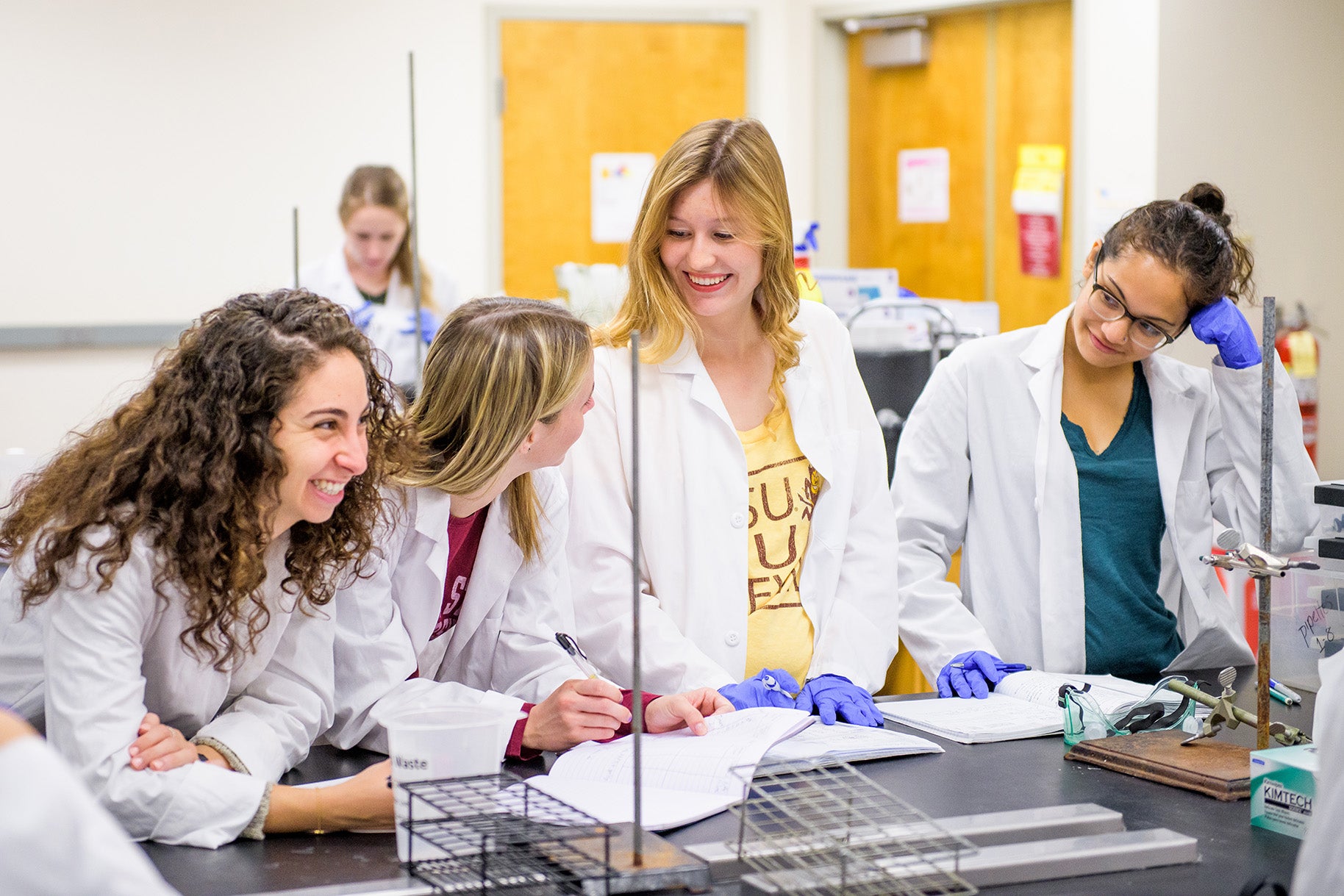 4 students in a lab setting wearing lab coats and laughing with each other.