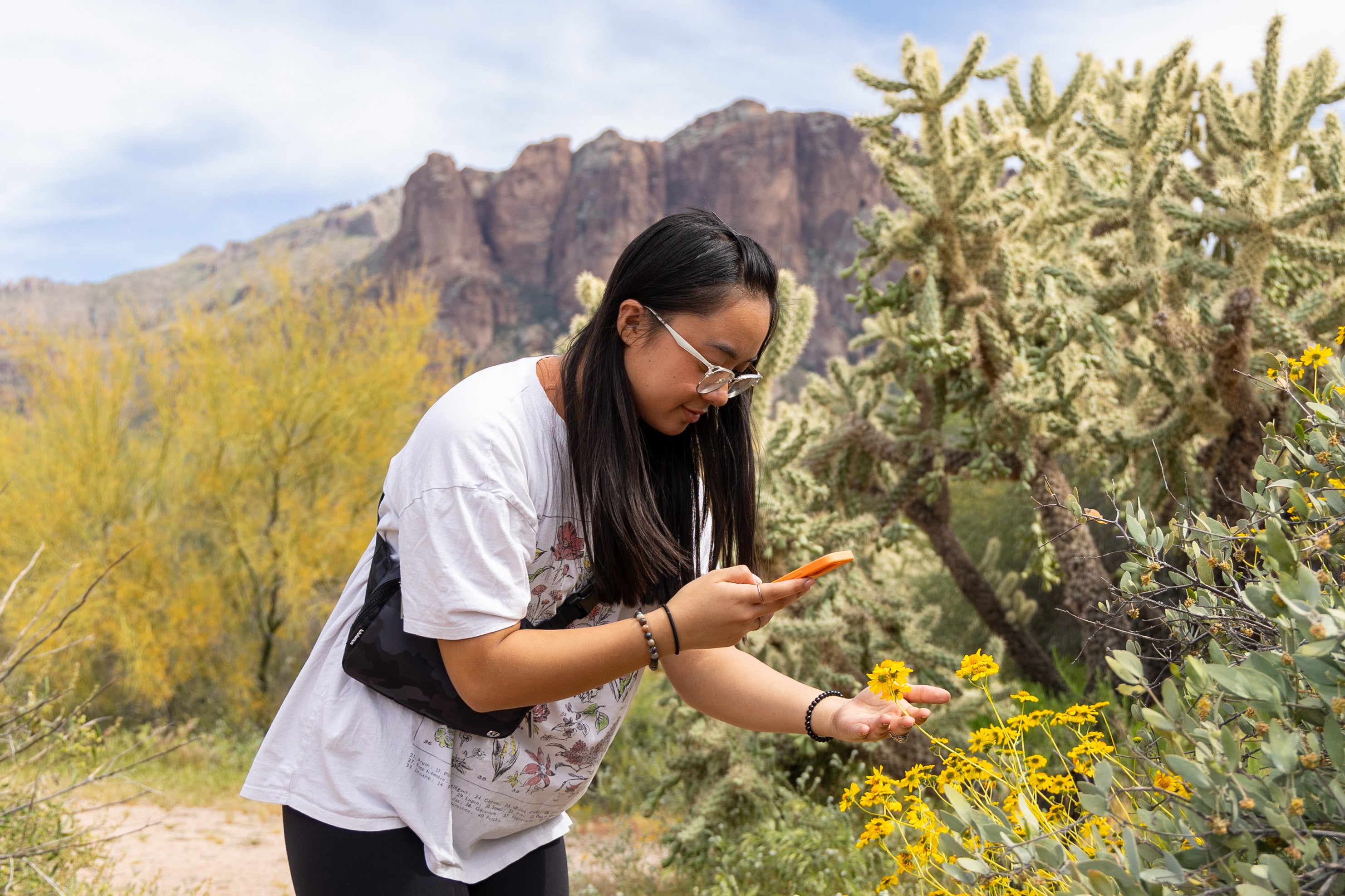 Woman standing outdoors amongst thick desert flora, inspecting a flower.