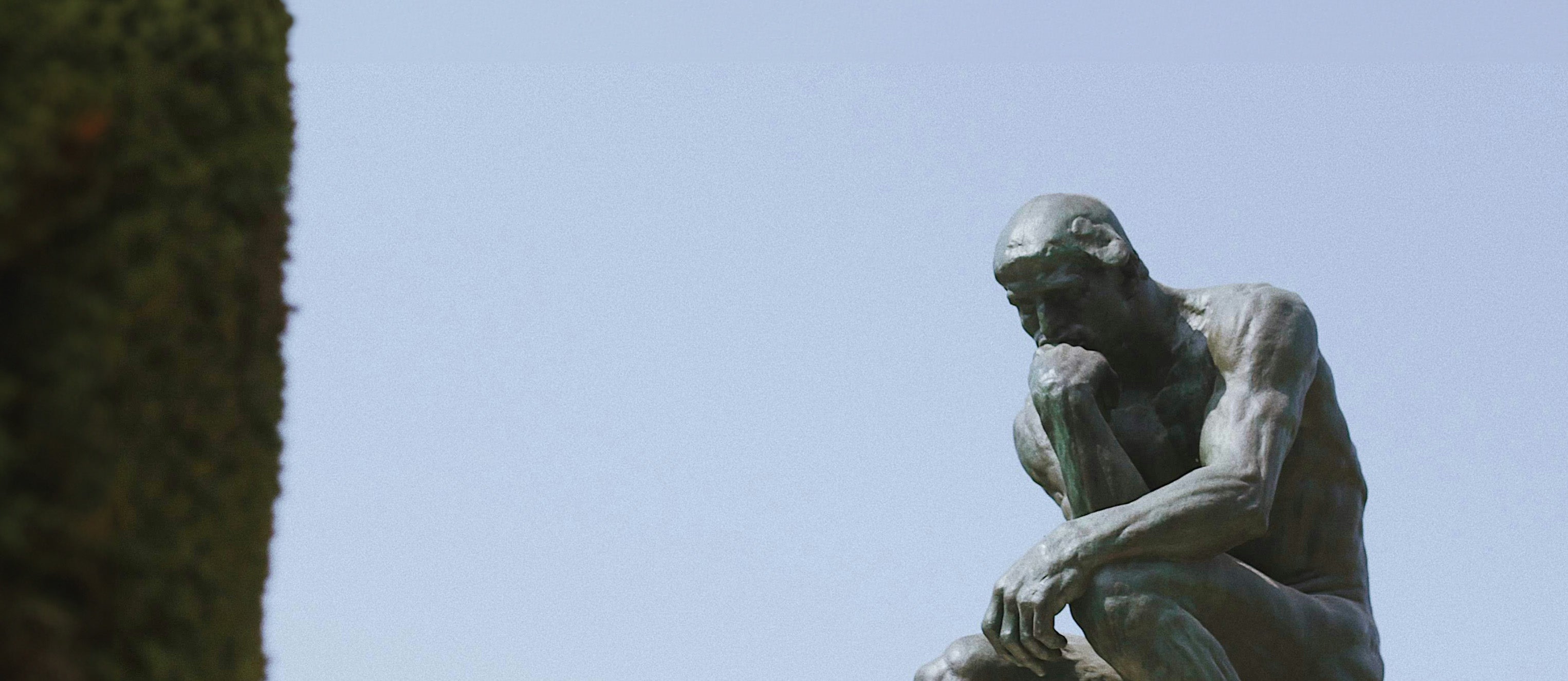 The Thinker statue in front of a blue sky with an evergreen shrub visible on the left side of the image
