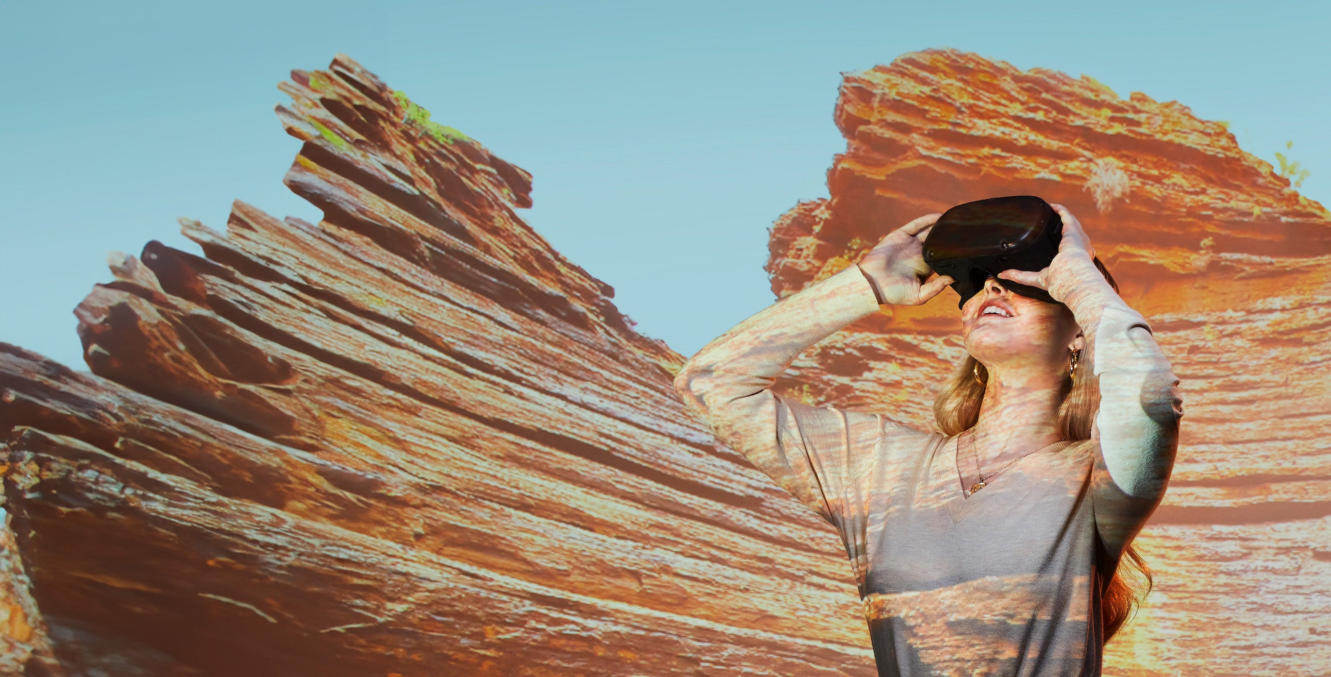 Woman with V-R goggles standing in front of a screen which has naturally red-colored cliffs projected on to it.