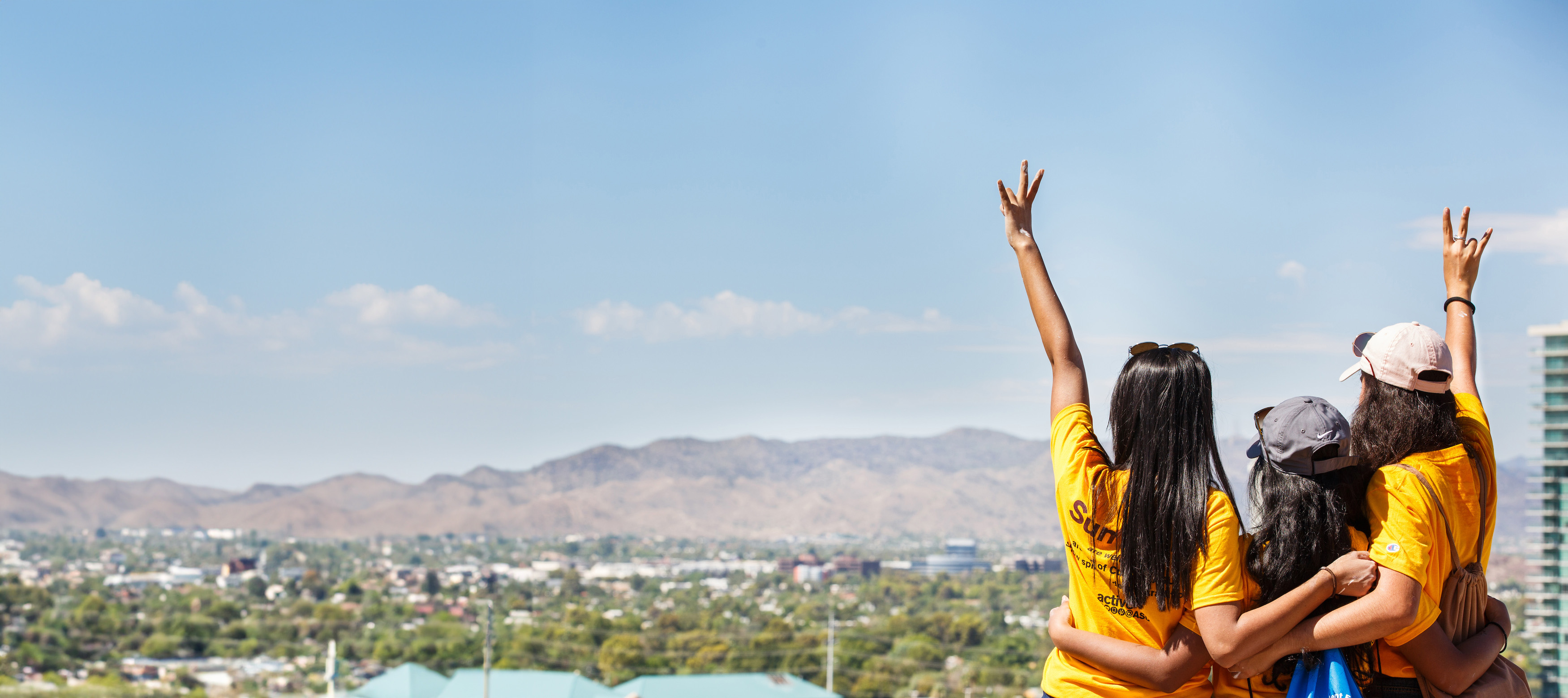 Three women wearing yellow with their backs turned facing a city-overlook and mountain range