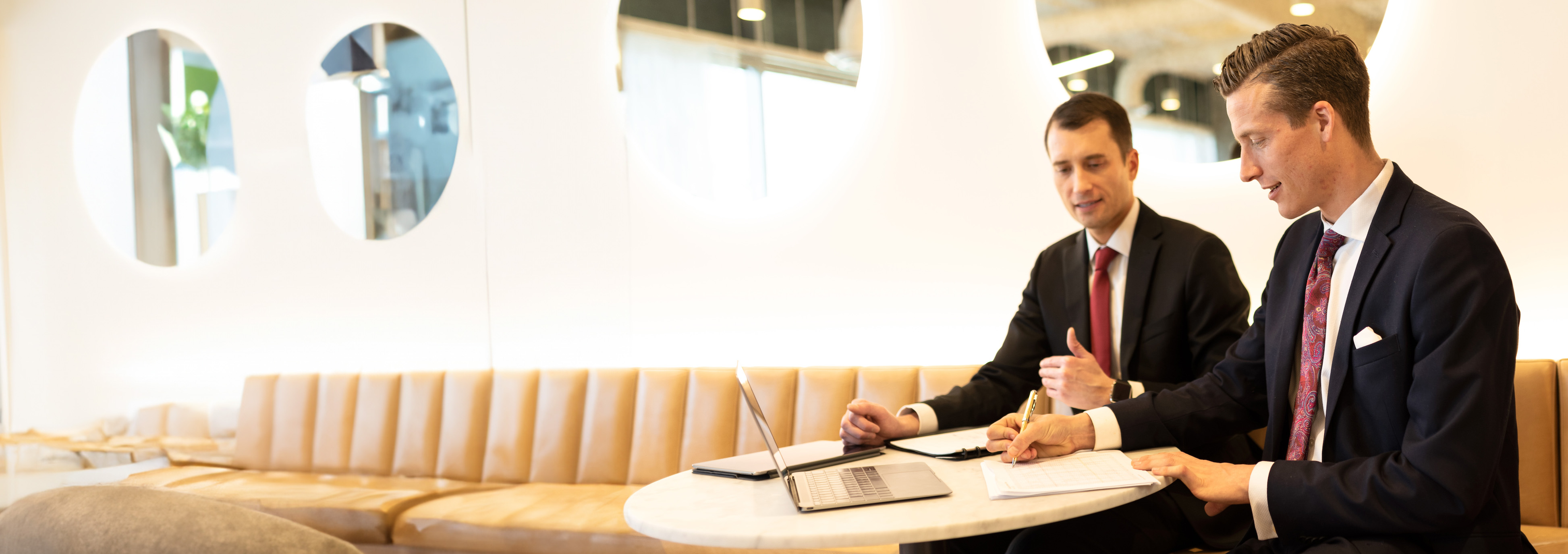 Two men sitting at a table in a white and beige room with their laptops and notebooks open