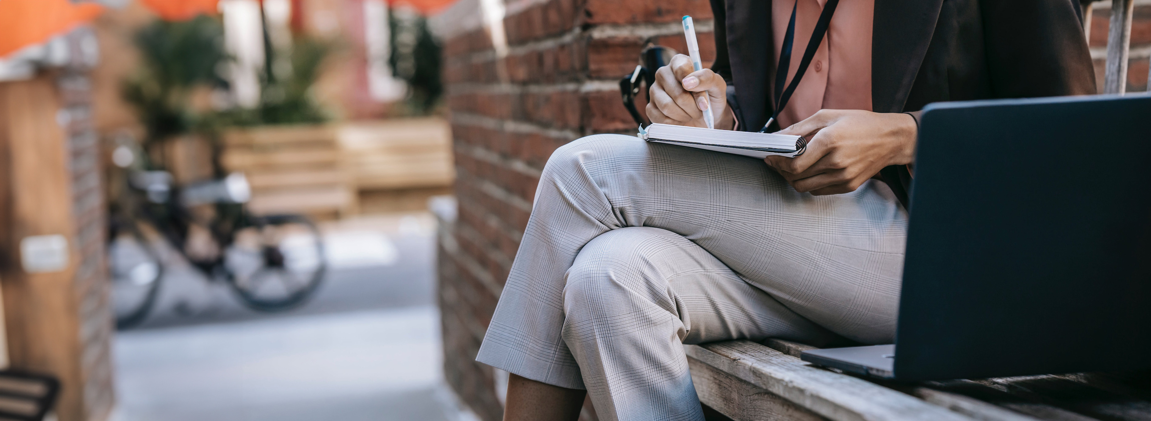 Woman with legs folded sitting on a bench in front of a brick wall holding a pen and notebook with a laptop sitting next to her