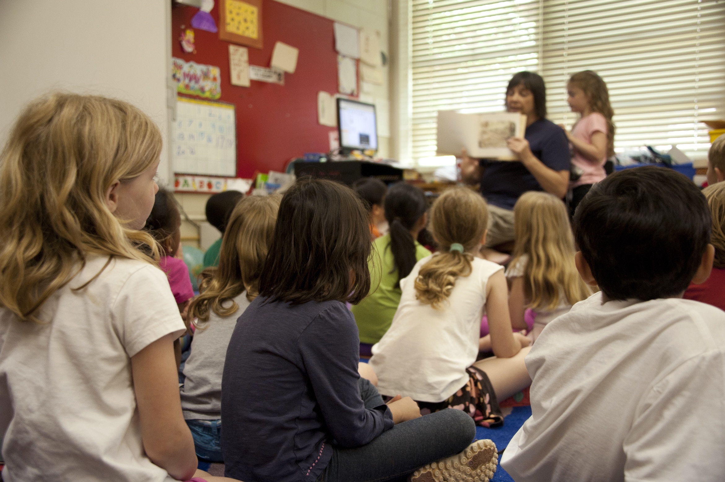 A teacher sitting in a chair at the front of a classroom full of kids sitting on the ground, reading the children a book.