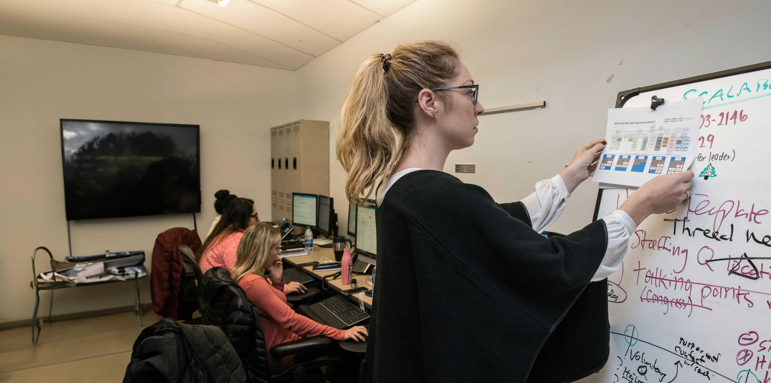 Small office with 3 women sitting at a shared desk on computers and one woman standing at a whiteboard with writing covering the entire board.