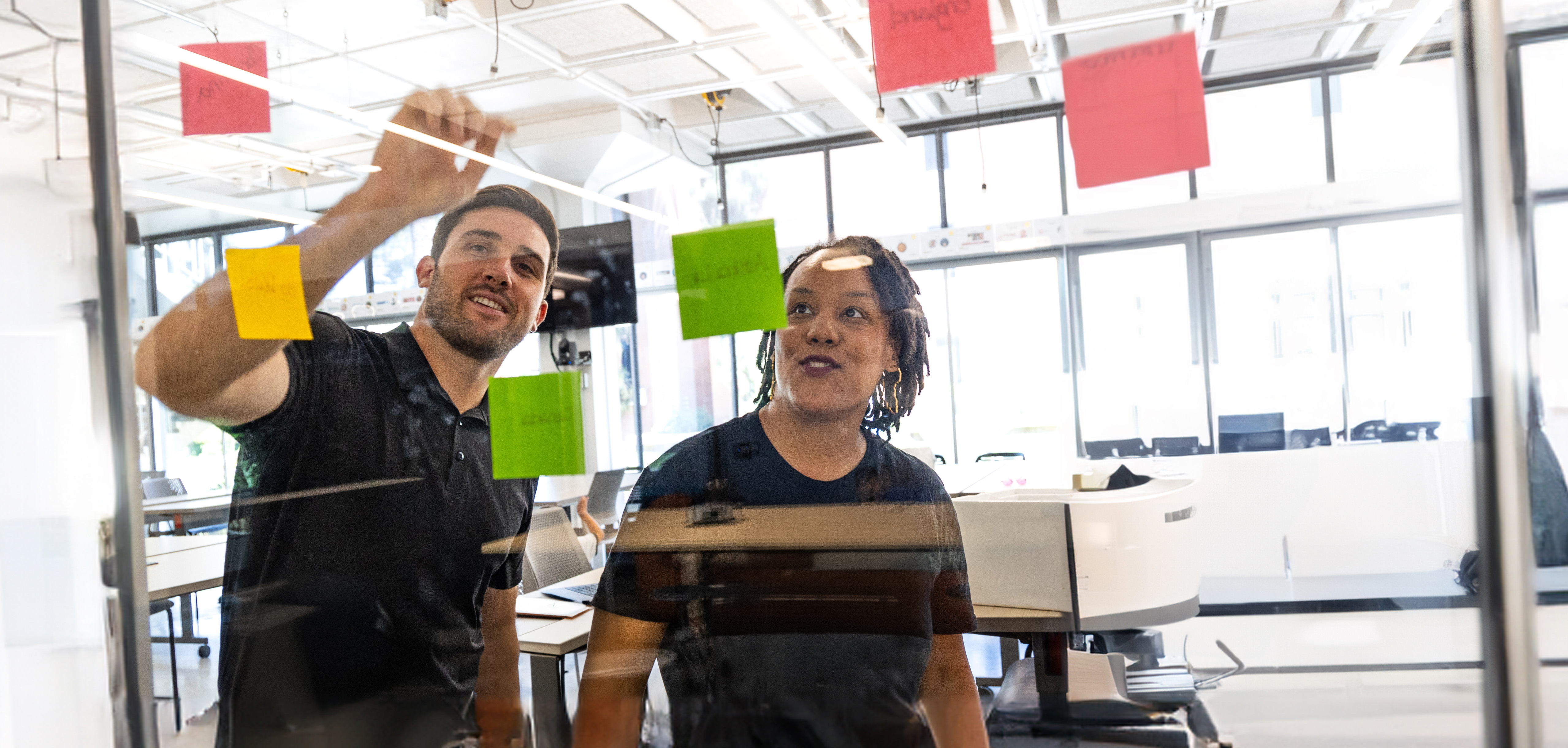 Two people standing in front of a clear glass wall, brainstorming with post-it notes
