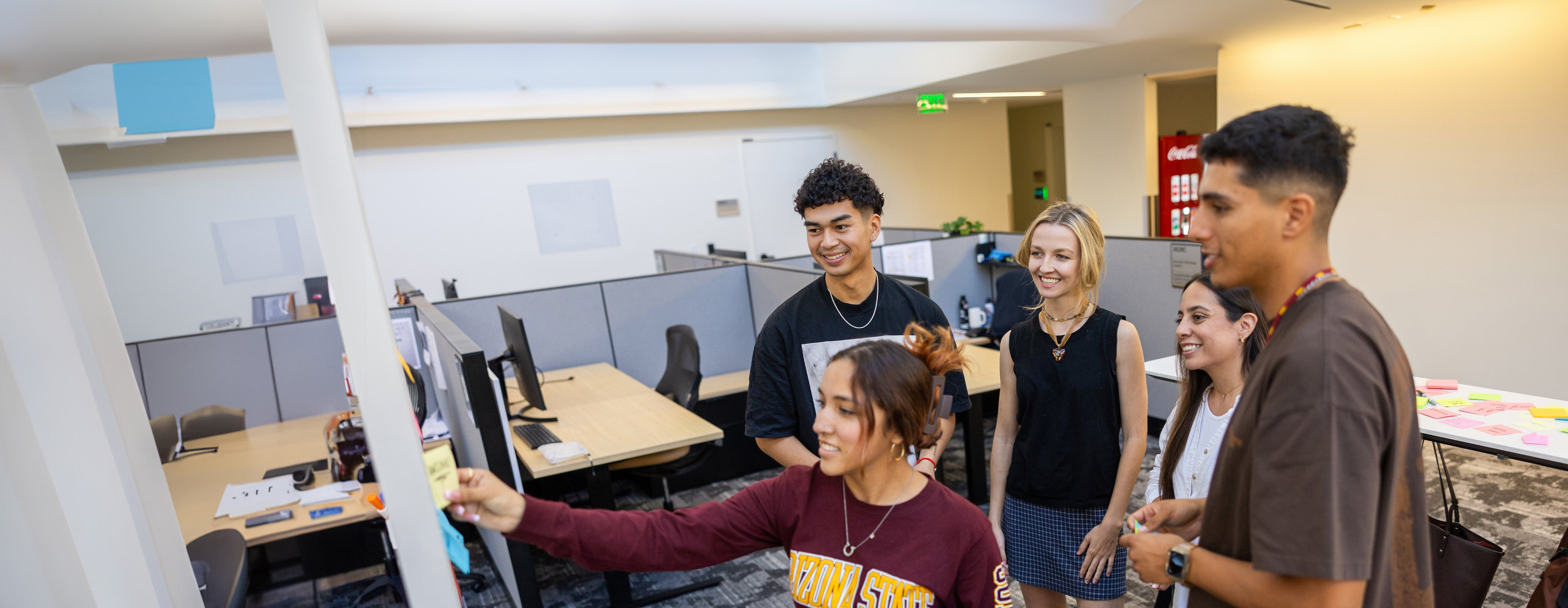 A group of five young adults collaborates and smiles in an office, with one placing a sticky note on a board.