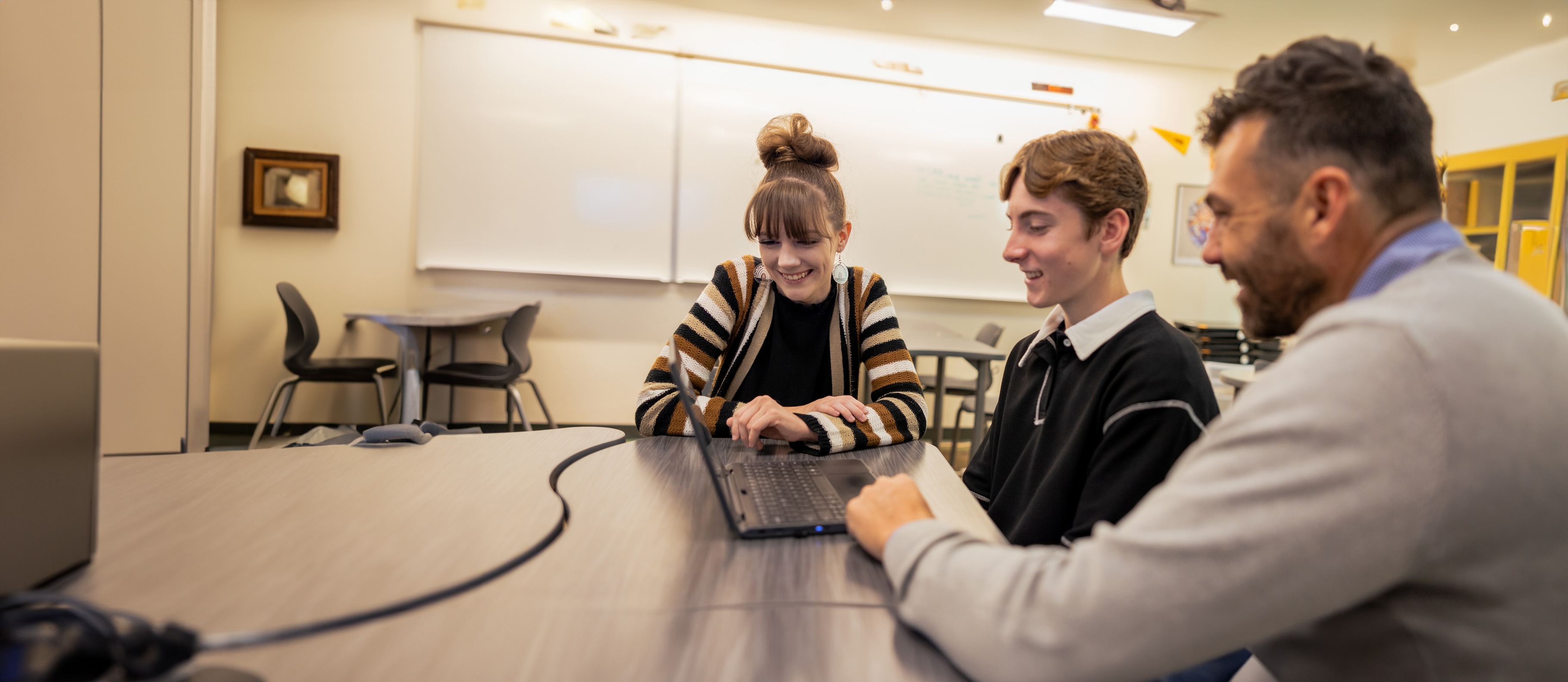 Three people, two students and an adult, looking at a laptop at a table in a classroom.