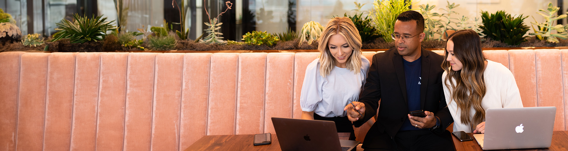 Three people sitting on a peach-colored cushioned bench with laptops, discussing work.