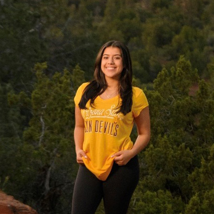 Nicole Irwin, a young woman wearing a gold A-S-U shirt standing in front of a forested backdrop.