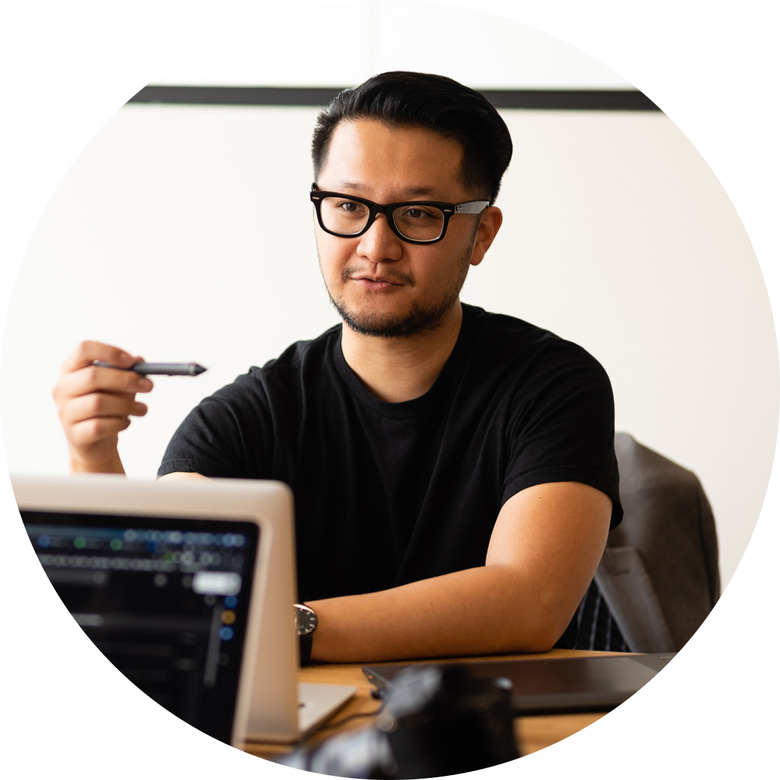 Man with slicked-back dark hair and wearing glasses, sitting at a table looking at a laptop and holding a pen