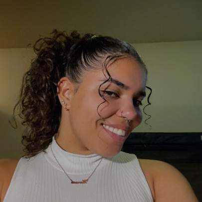 Nevaeh Shank, a young woman with curly dark hair tied back, wearing a white sleeveless top and a necklace and smiling.